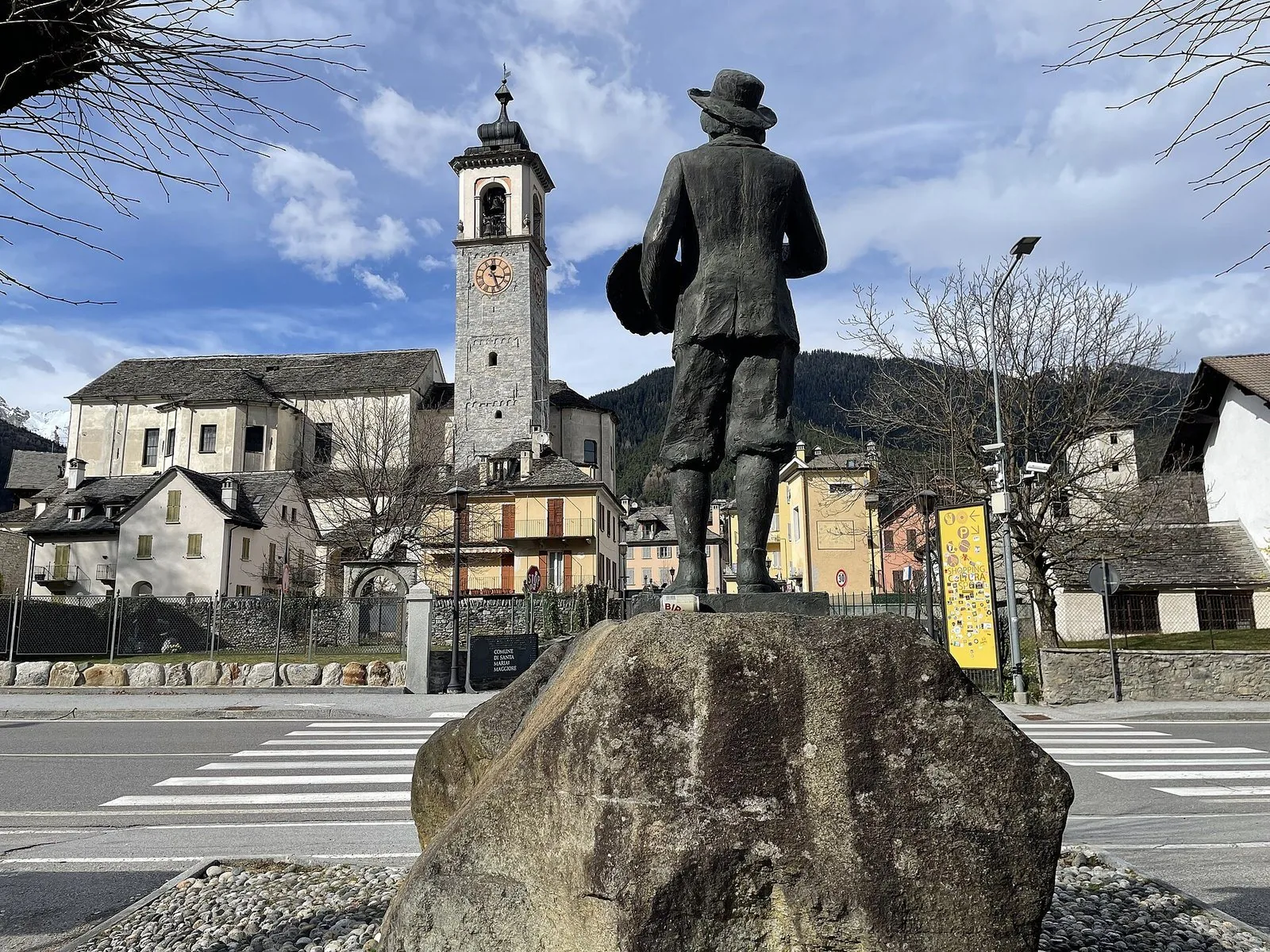 La chiesa di Santa Maria Assunta con il campanile in pietra a Santa Maria Maggiore, vista oltre una statua in bronzo su un masso