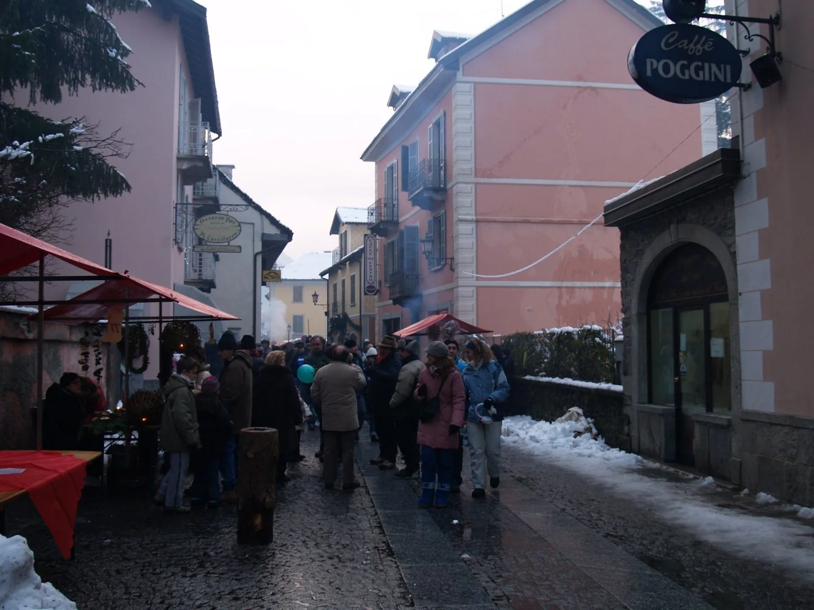 Una via del centro di Santa Maria Maggiore durante il mercatino di Natale, con bancarelle rosse, visitatori e neve ai bordi della strada